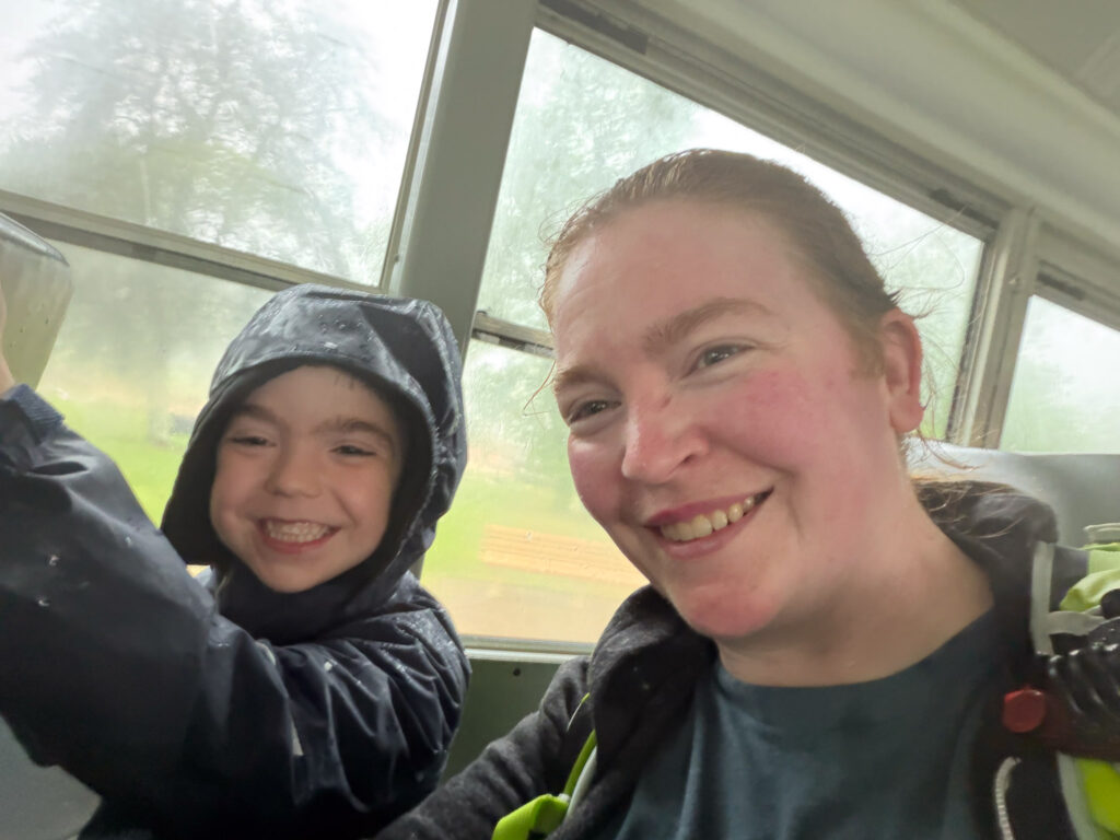 A woman and child smile together after getting soaked in the rain during the Domes and Dripstones Tour at Mammoth Cave, wet trees and ground visible through large windows behind them.