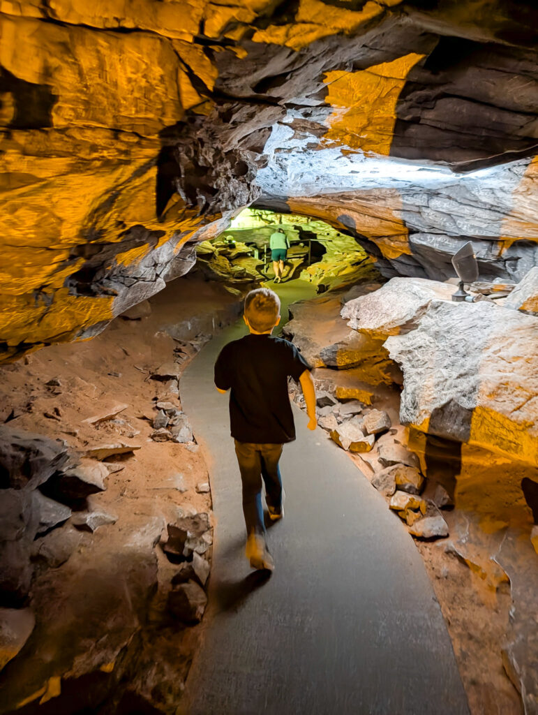 A child walks along a paved path through a narrow limestone passage on Mammoth Cave's Historic Tour, with warm orange lighting illuminating the rocky walls and a larger chamber visible ahead.