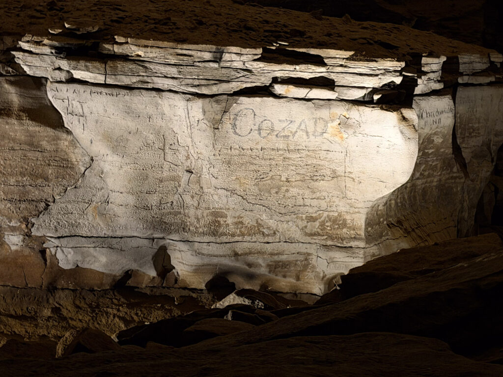 A limestone wall in Mammoth Cave bearing the carved name 'Cozad' and other faint historical inscriptions, illuminated by artificial light during the Historic Tour, with layered sedimentary ceiling above and rubble on the cave floor.