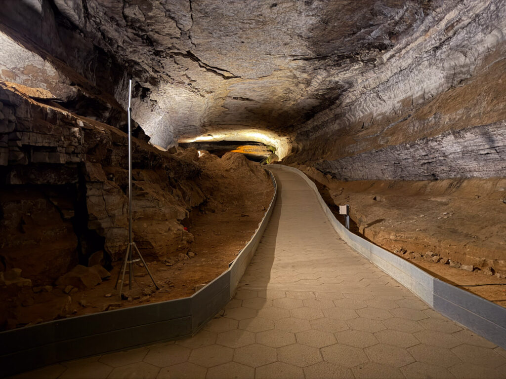 Paved walkway curving through a broad limestone passage on Mammoth Cave's Historic Tour, with artificial lighting illuminating the textured gray ceiling and rocky formations along both sides.