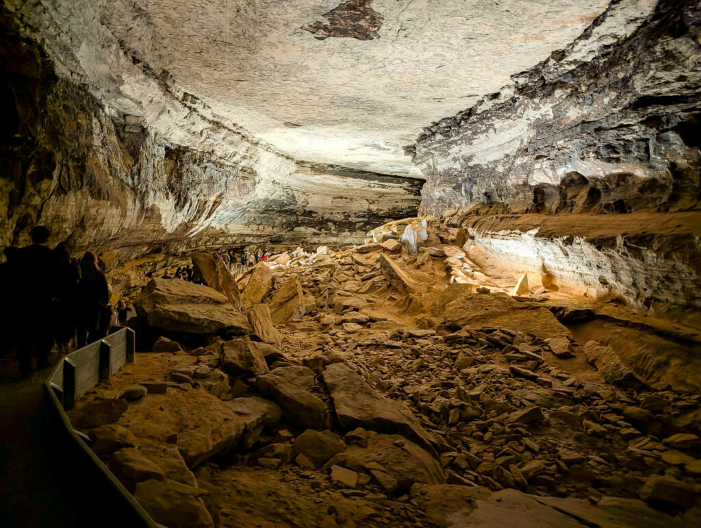 A guided Historic Tour group walks a railed path through a vast Mammoth Cave chamber, its vaulted white limestone ceiling and boulder-strewn floor emphasizing the cavern's immense scale.