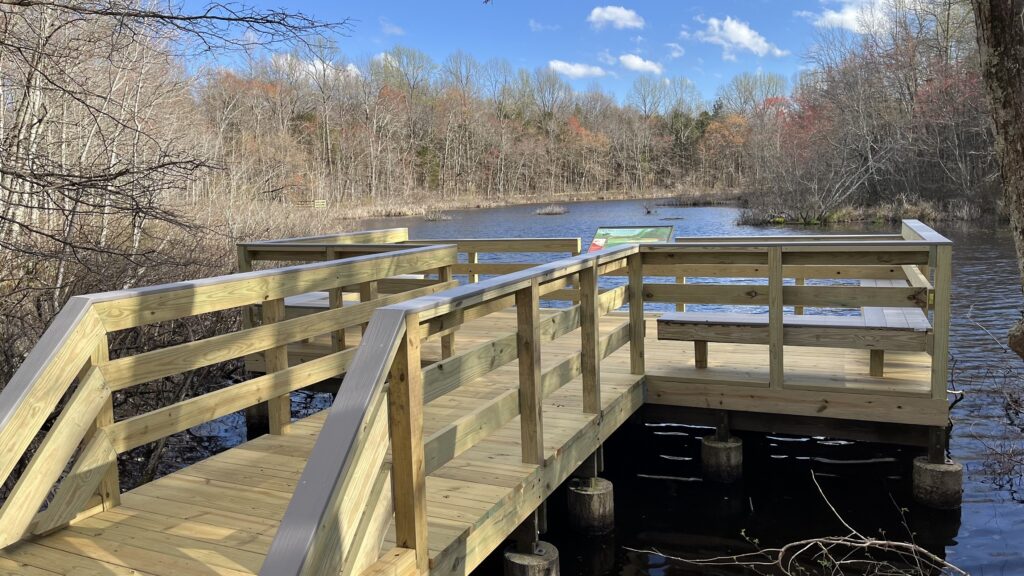 A new light-wood accessible observation deck with horizontal railing extends over the still dark water of Sloan's Crossing Pond at Mammoth Cave, surrounded by autumn trees under a partly cloudy sky.