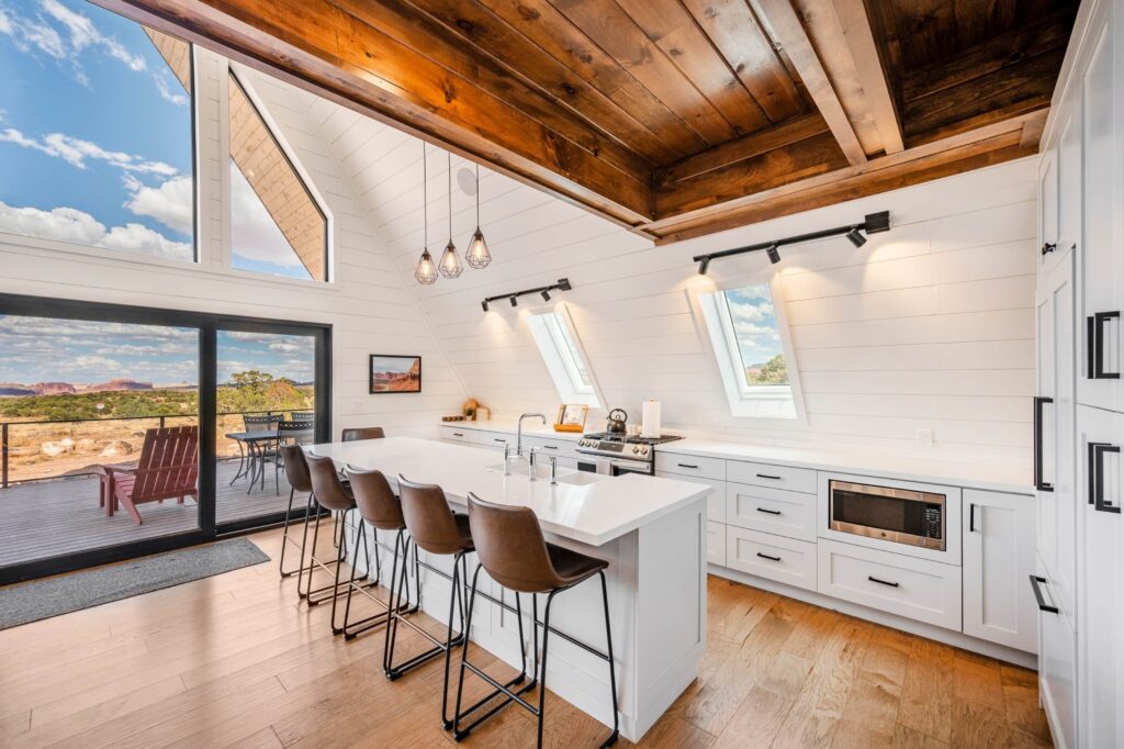 A-frame cabin kitchen at Cabins at Capitol Reef in Torrey, UT, with white shiplap walls, leather barstools at a quartz island, and a sliding glass door opening to red rock desert views.