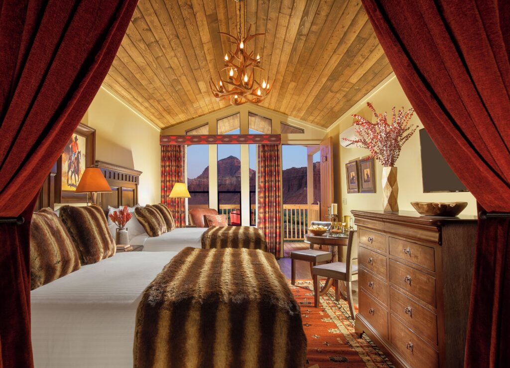Western-themed guest room at Capitol Reef Resort with two beds, faux-fur throws, antler chandelier, and large windows overlooking red rock canyon formations at dusk.