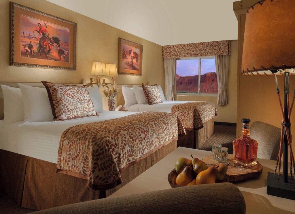 Western-themed double-queen room at Capitol Reef Resort in Torrey, UT, with earth-tone bedding, antler lamp, cowboy art, and a window view of red sandstone cliffs at dusk.