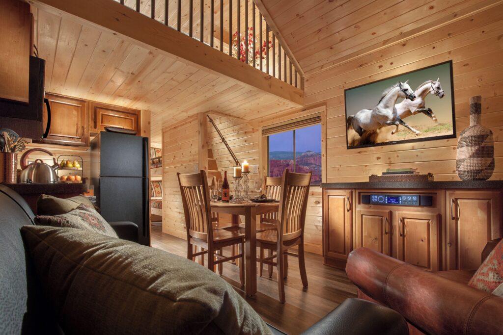 Cozy log cabin interior at Capitol Reef Resort in Torrey, UT, with knotty pine walls, a loft, living and dining areas, compact kitchen, and a window framing red rock formations outside.