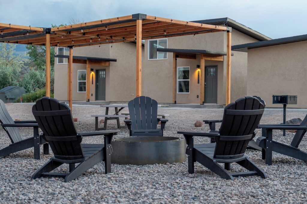 Gravel courtyard at Casitas at Capitol Reef in Torrey, UT, with a wood-beam pergola strung with globe lights, four Adirondack chairs around a fire pit, and two casita entrances glowing with wall sconces at dusk.