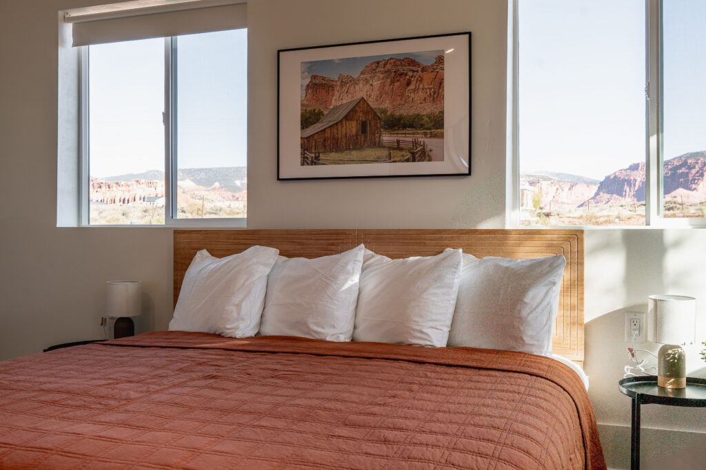 Casitas at Capitol Reef guest room bed with terracotta quilt, flanked by windows revealing red sandstone formations under a bright blue sky in Torrey, UT.
