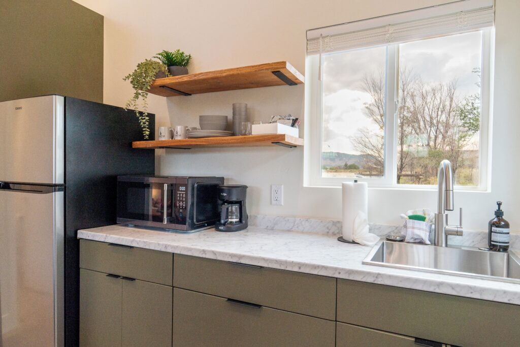 Kitchen in a Casitas at Capitol Reef rental in Torrey, UT, featuring olive-green cabinets, marble-look countertops, floating wood shelves with dishware, and a window overlooking bare trees and low hills.