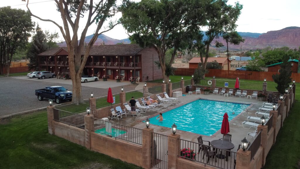Elevated dusk view of the Chuckwagon Lodge in Torrey, UT, showing a turquoise outdoor pool with guests relaxing poolside, a two-story log-style motel building with balconies, and red sandstone cliffs in the background.