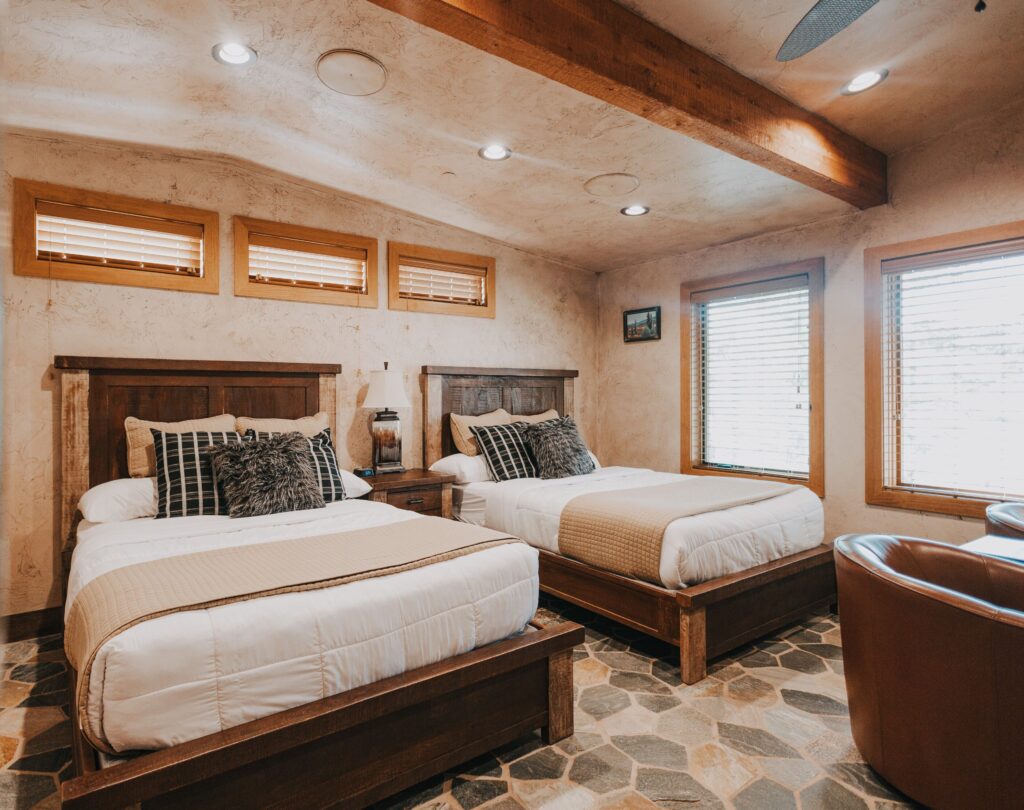 Guest room at Cougar Ridge Resort in Torrey, UT with two queen beds featuring dark wood frames and white bedding, stone tile floors, exposed ceiling beams, and plaster walls with natural light from side windows.