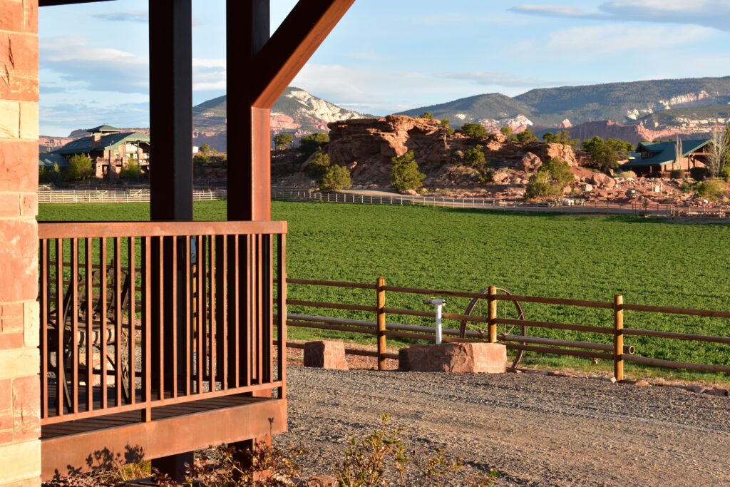 Covered porch at Cougar Ridge Resort in Torrey, UT, overlooking a gravel path, green field, and ranch buildings beneath dramatic red rock formations and pinyon-juniper hills near Capitol Reef.
