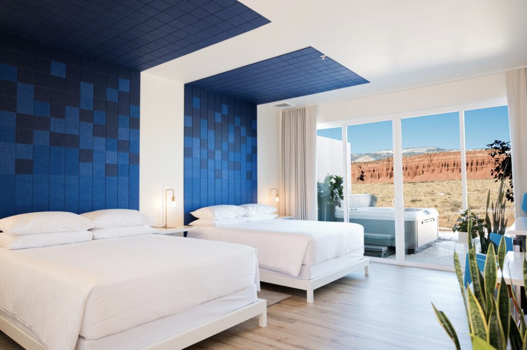 Skyview Hotel guest room in Torrey, UT featuring two platform beds against a blue tiled accent wall, with floor-to-ceiling glass doors opening to a private patio hot tub overlooking red rock canyon views.