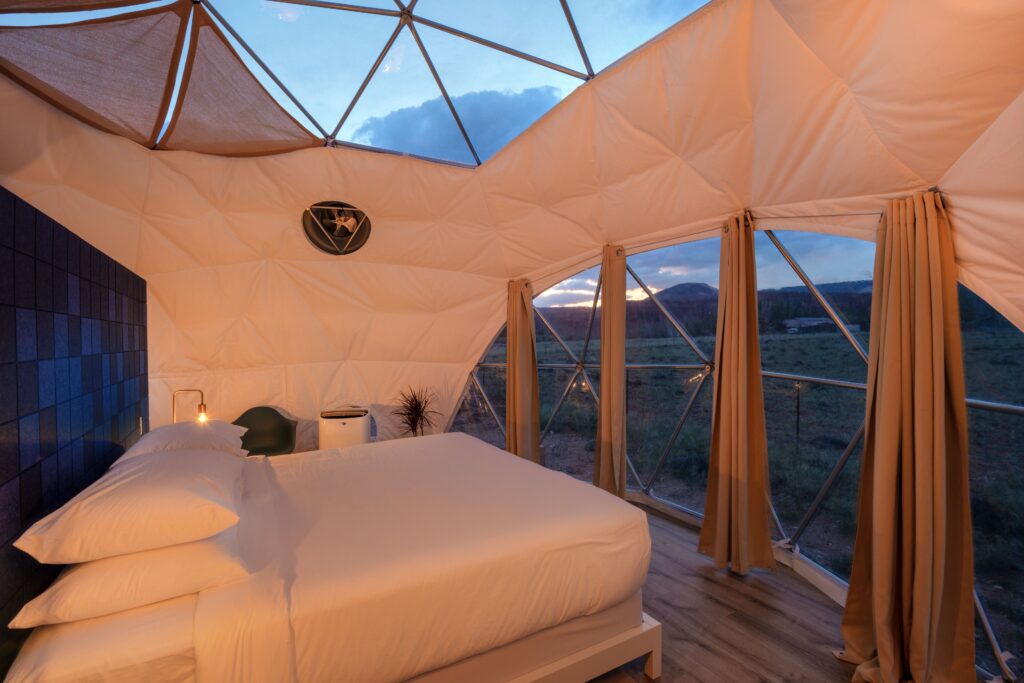Interior of a geodesic glamping dome at Skyview Hotel in Torrey, UT, with a made bed, warm sconce lighting, and a curved glass wall framing a twilight view of rolling hills.