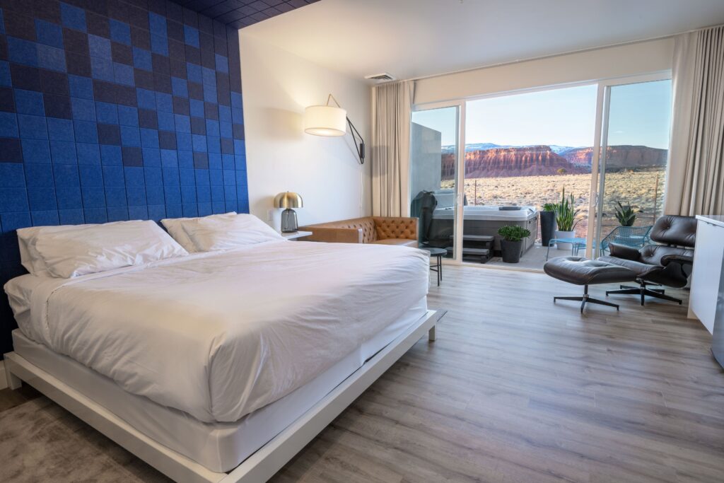 Skyview Hotel guest room in Torrey, UT featuring a king bed with a bold blue checkered accent wall, light wood flooring, and floor-to-ceiling sliding doors opening to a private balcony hot tub with sweeping red sandstone cliff views.