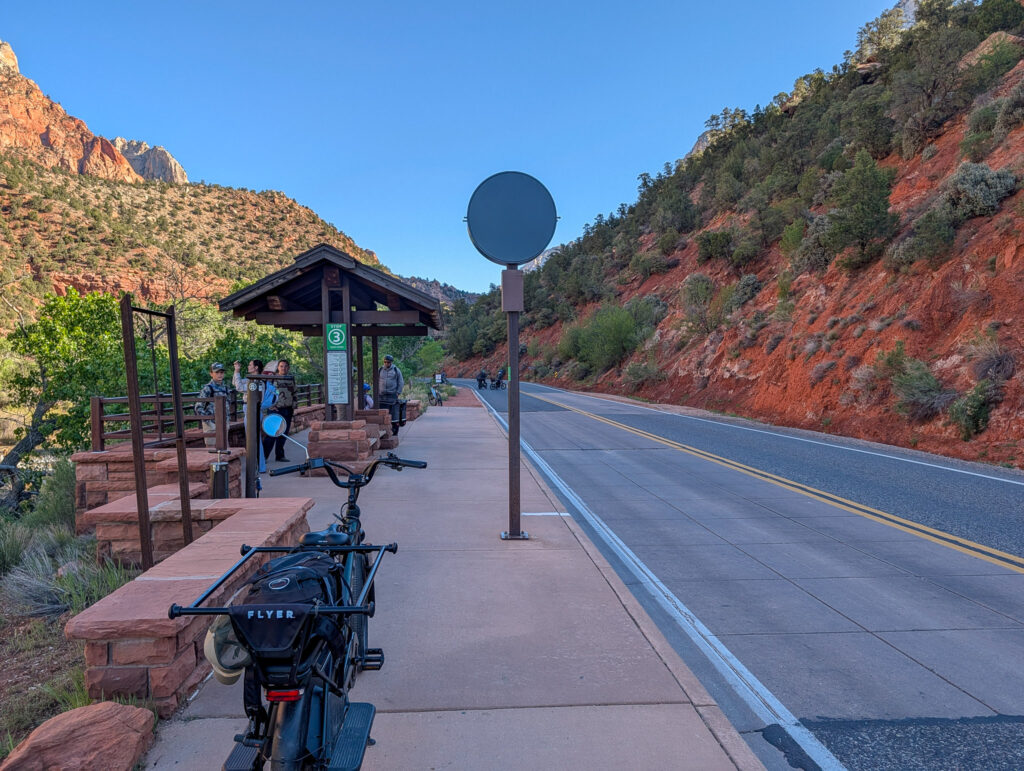 Visitors wait for the Zion Canyon Line shuttle. A rad wagon e-bike sits parked along the side of the sidewalk, where the Pa'rus trail exits from below the road.