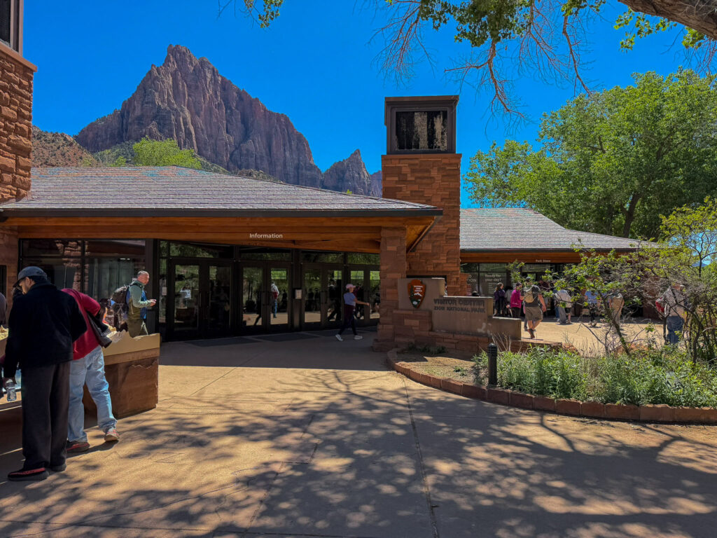 Visitors mill near the wood-and-stone Zion Canyon Visitor Center entrance with the Watchman peak rising sharply behind it.